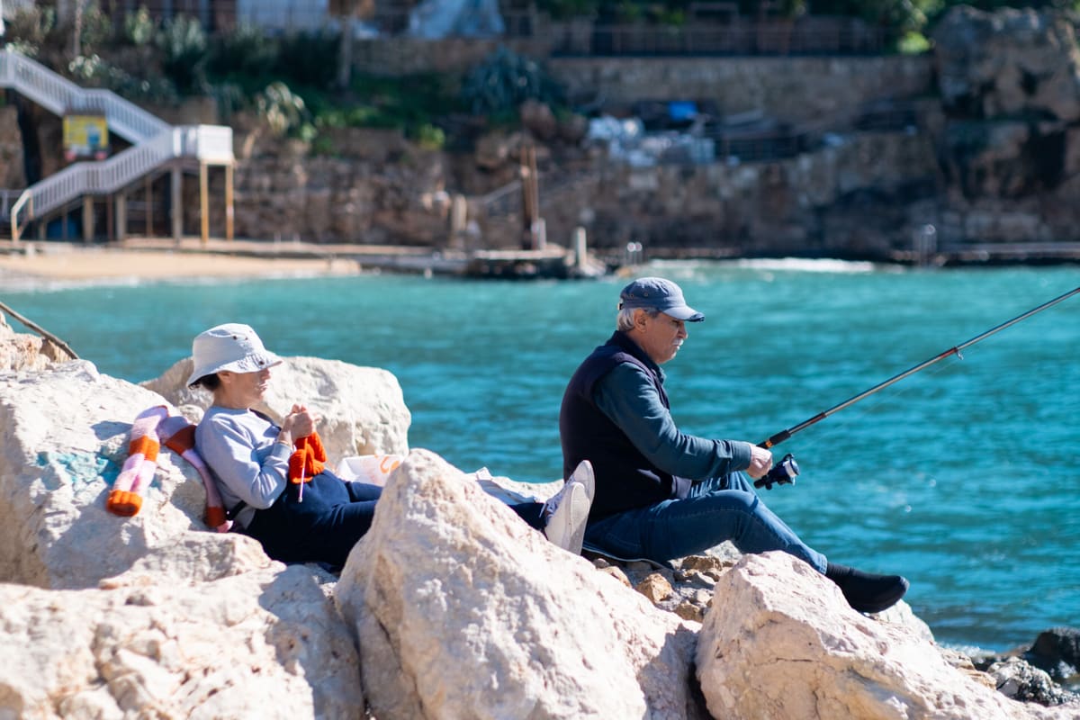 A couple on coastal rocks, the man fishing while the woman knits beside clear water